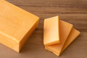 A block of cheddar cheese with three slices placed beside it on a wooden surface