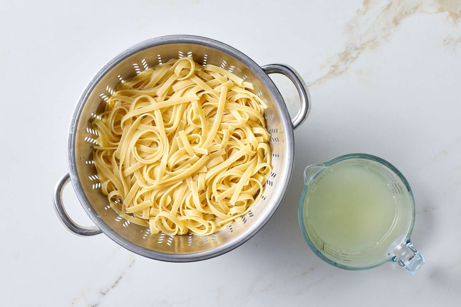 Cooked fettuccine draining in a colander next to a measuring cup of pasta water