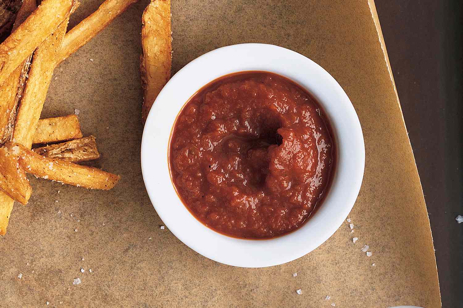 A dish of ketchup accompanied by French fries on parchment paper