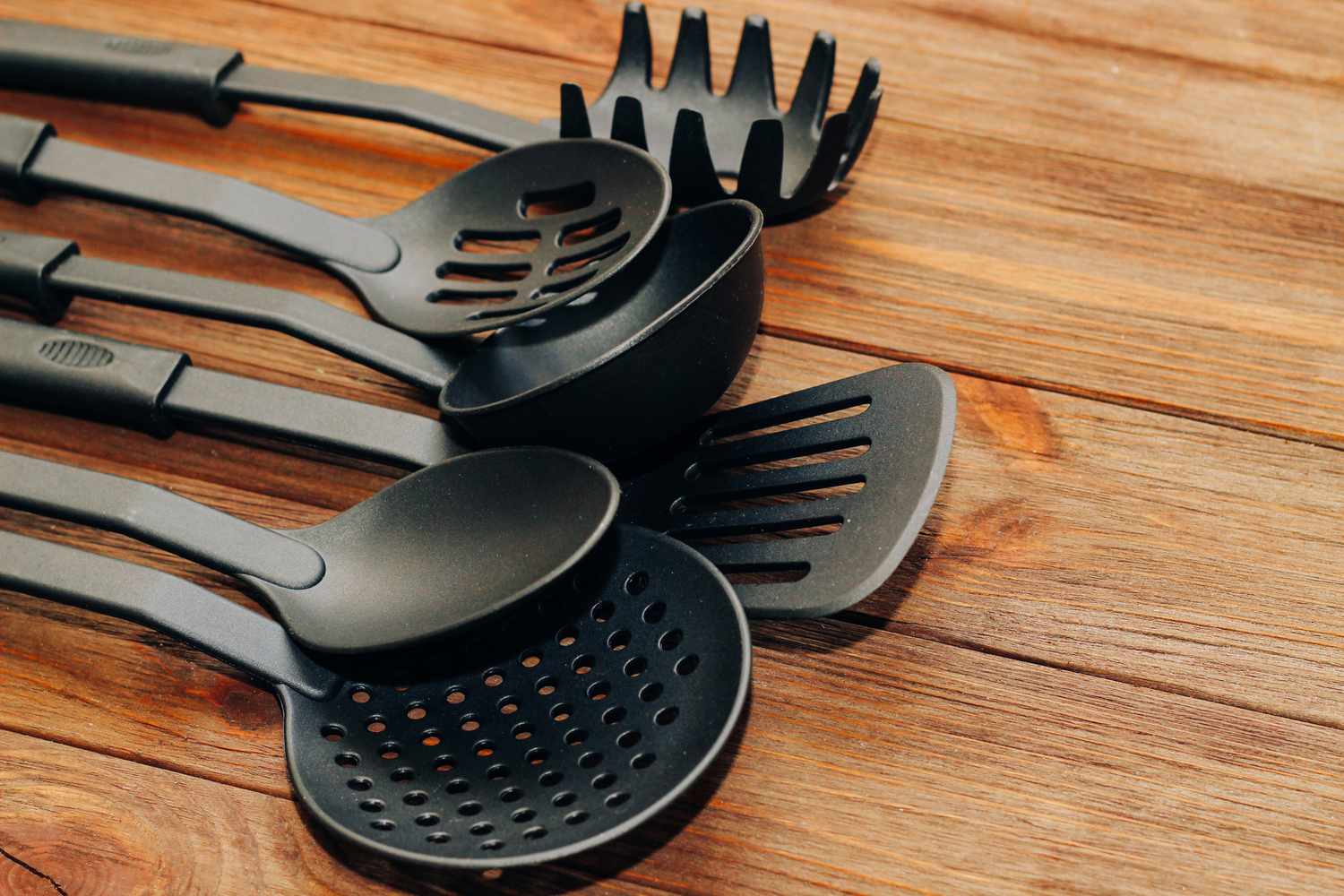 A set of black kitchen utensils including ladles and a pasta server laid on a wooden surface