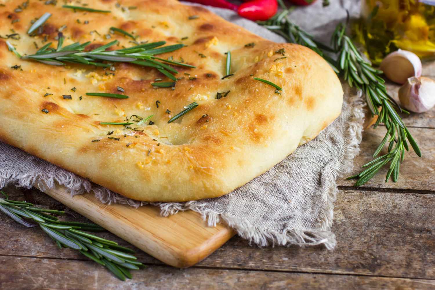 A piece of focaccia bread garnished with rosemary placed on a wooden board over a cloth with additional rosemary sprigs nearby
