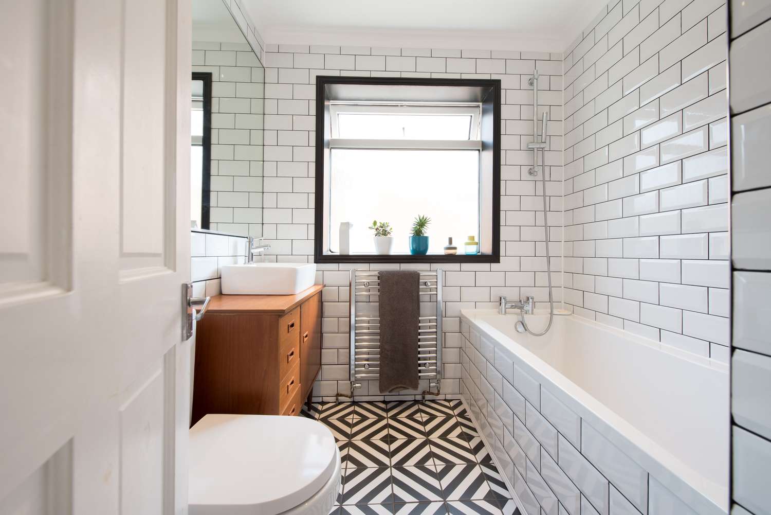 Modern bathroom with a bathtub white subway tiles geometric floor tiles wood vanity and a central window with plants on the sill