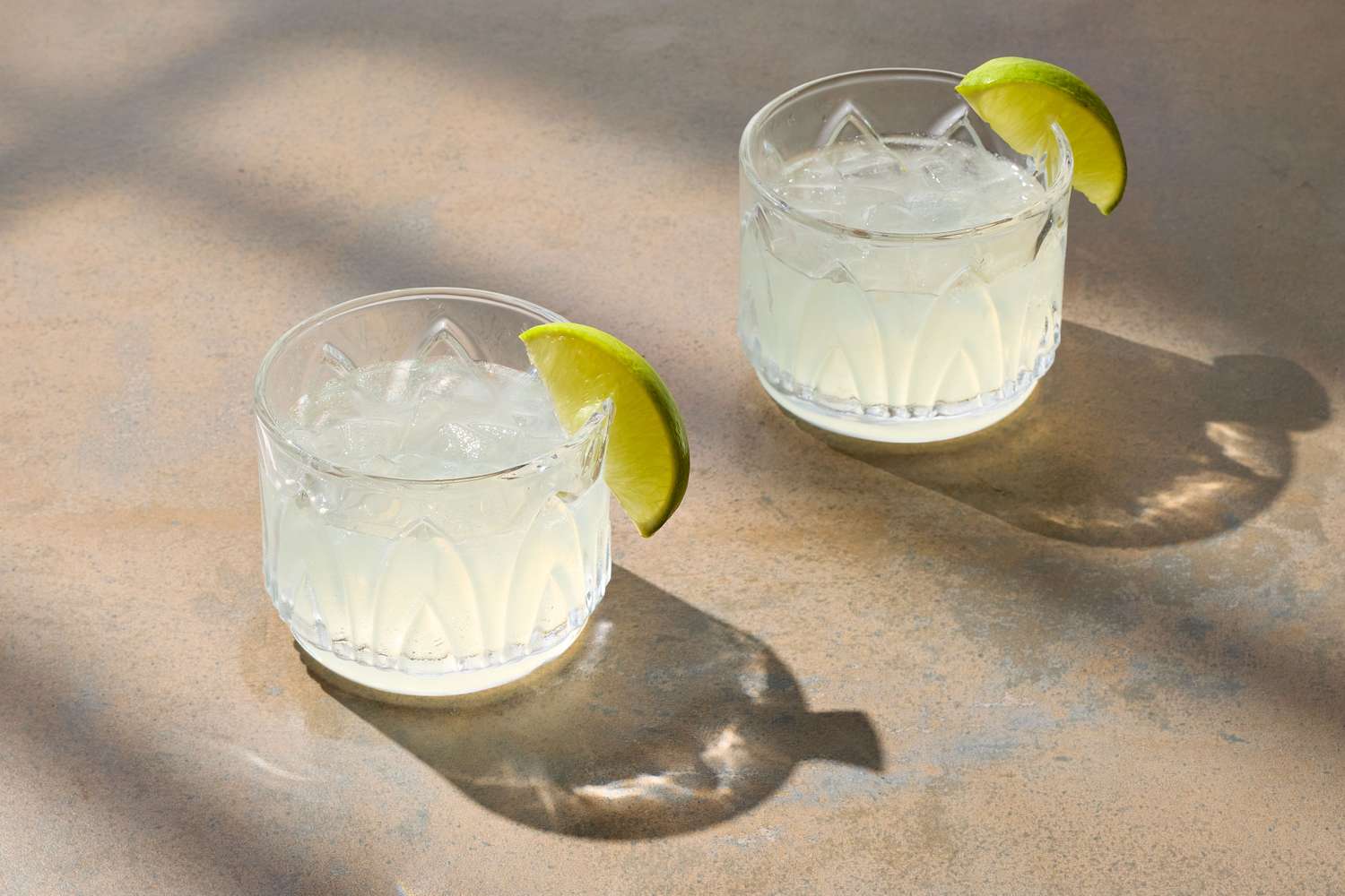 Two gin gimlet cocktails garnished with lime wedges on a surface with light and shadow patterns