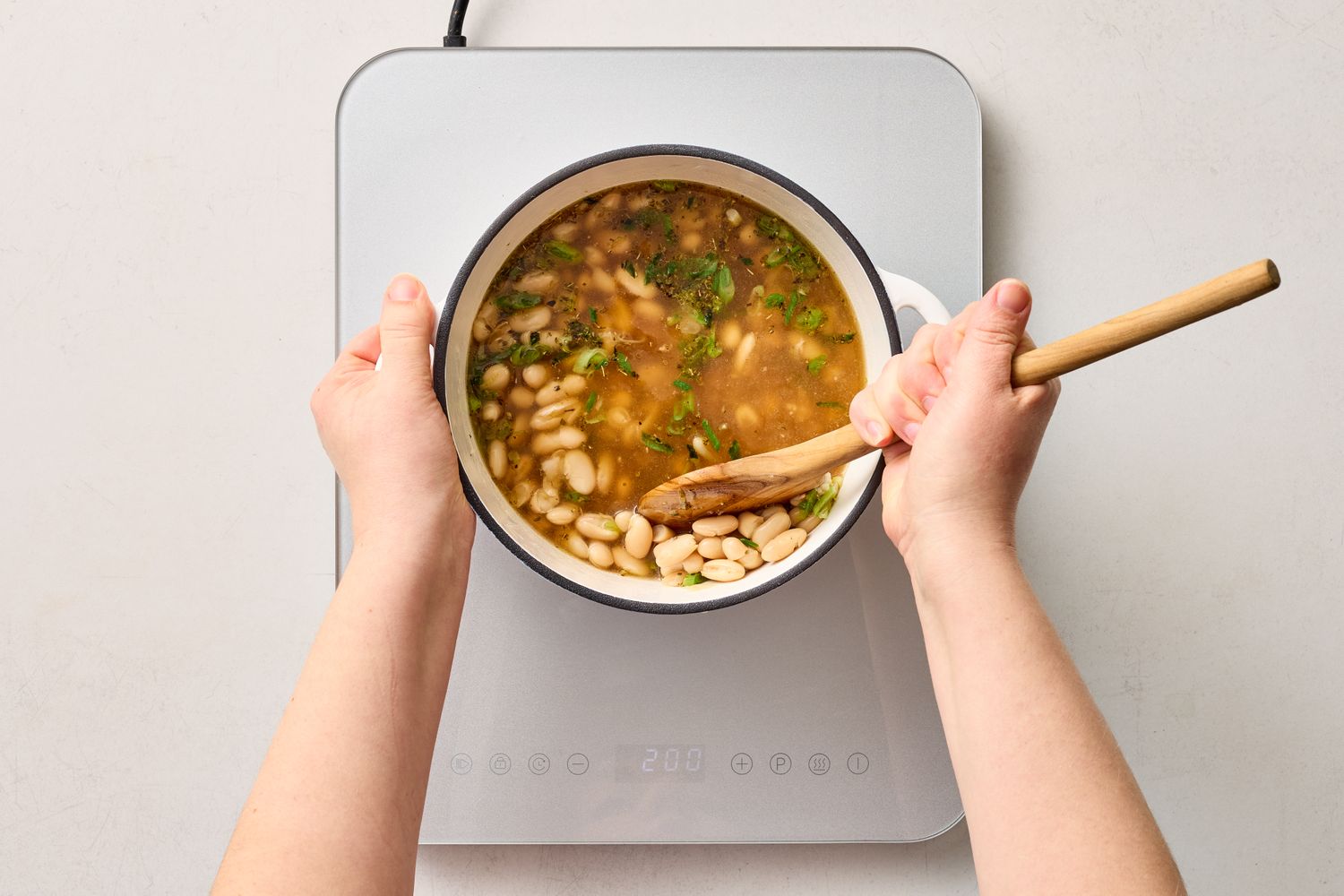 A pot of bean soup being stirred with a wooden spoon on a stovetop garnished with herbs