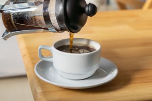 French press pouring coffee into a white cup on a saucer on a table