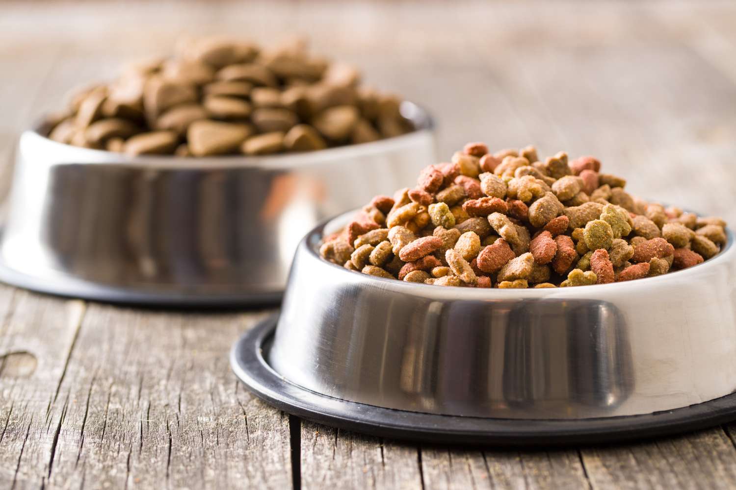Two stainless steel bowls on a wooden surface one containing dry dog food and the other containing kibble