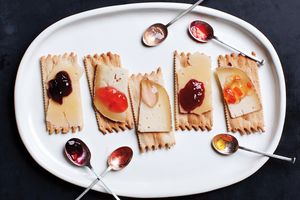 Crackers topped with slices of cheese and various fruit preserves arranged on a white plate with accompanying spoons of jam