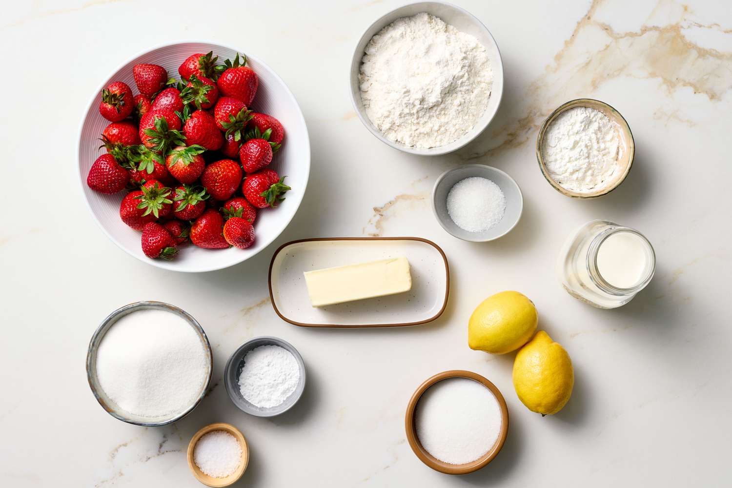 Ingredients for strawberry cobbler displayed on a surface, including strawberries, flour, sugar, butter, lemon, and milk