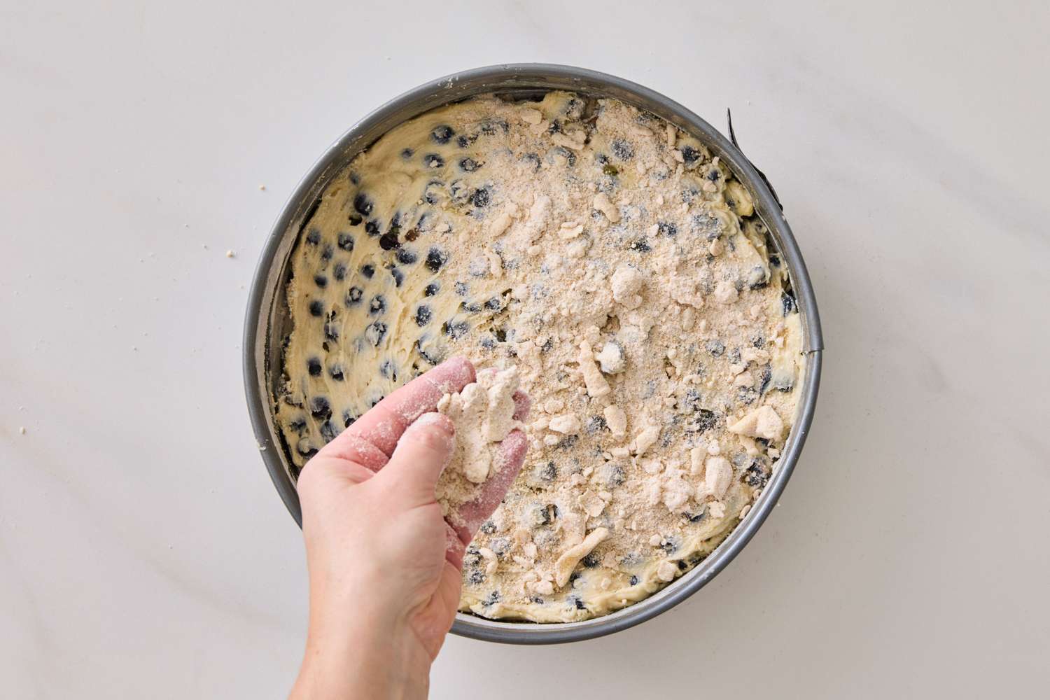 Person adding crumb topping to a blueberry dessert in a round baking pan