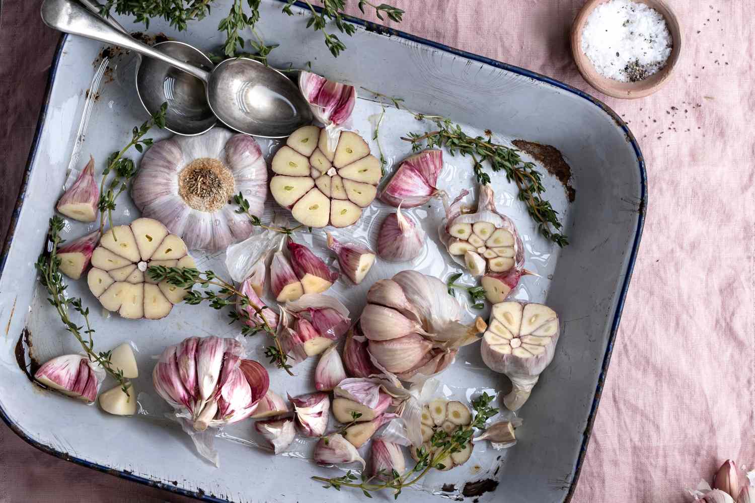 Halved garlic bulbs and sprigs of thyme in a baking dish with spoons on the side