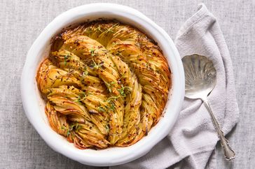A roasted potato dish baked in a circular pattern in a white dish garnished with herbs served with a metal spoon on the side