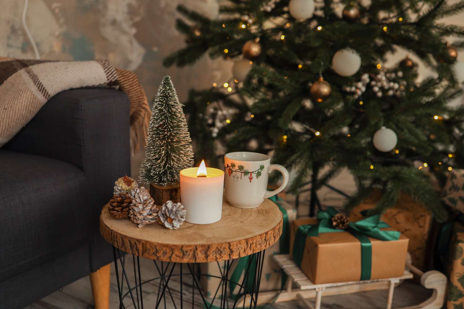 Christmasthemed decor setup with a table holding a candle mini tree and cup near a Christmas tree with ornaments and gifts underneath