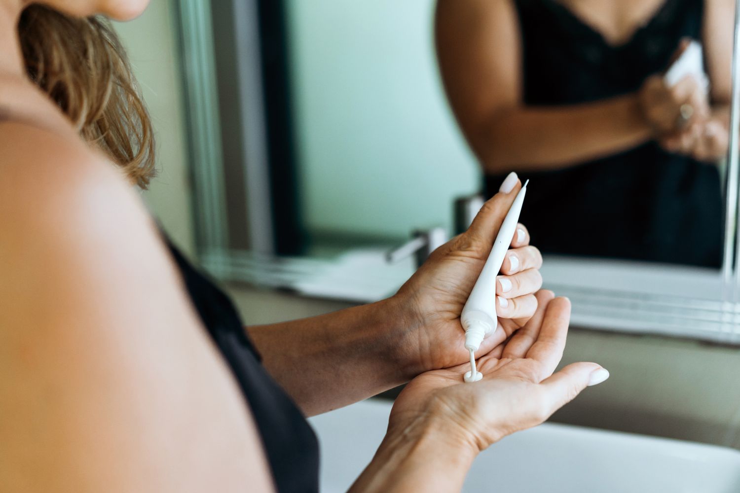Person squeezing lotion from a tube onto their hand by a sink