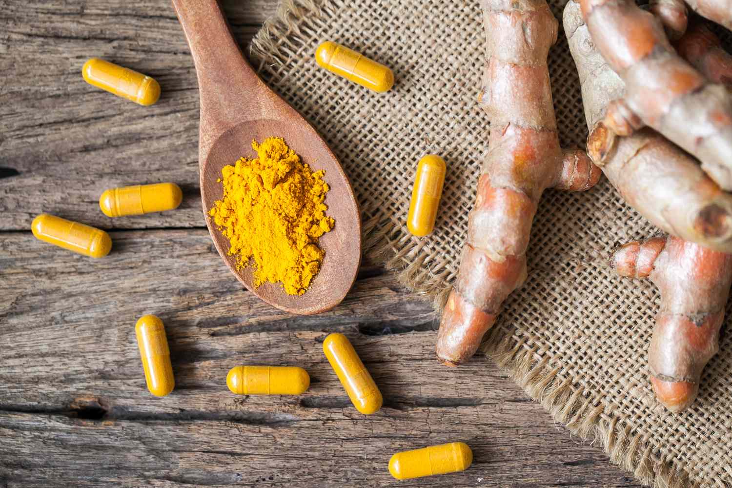 Turmeric powder in a wooden spoon surrounded by capsules and turmeric roots on a rustic surface