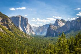 A natural landscape of a forested valley with prominent rocky mountain cliffs and a waterfall in the distance