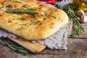 A piece of focaccia bread garnished with rosemary placed on a wooden board over a cloth with additional rosemary sprigs nearby