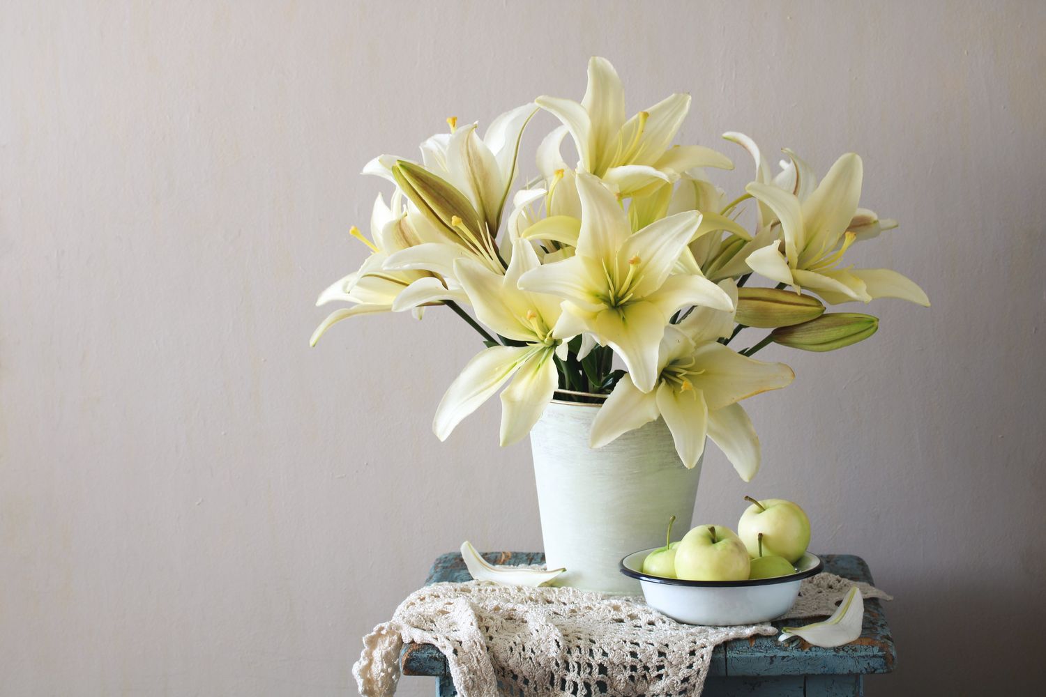 White lilies in a vase on a table with a lace cloth and a bowl of apples
