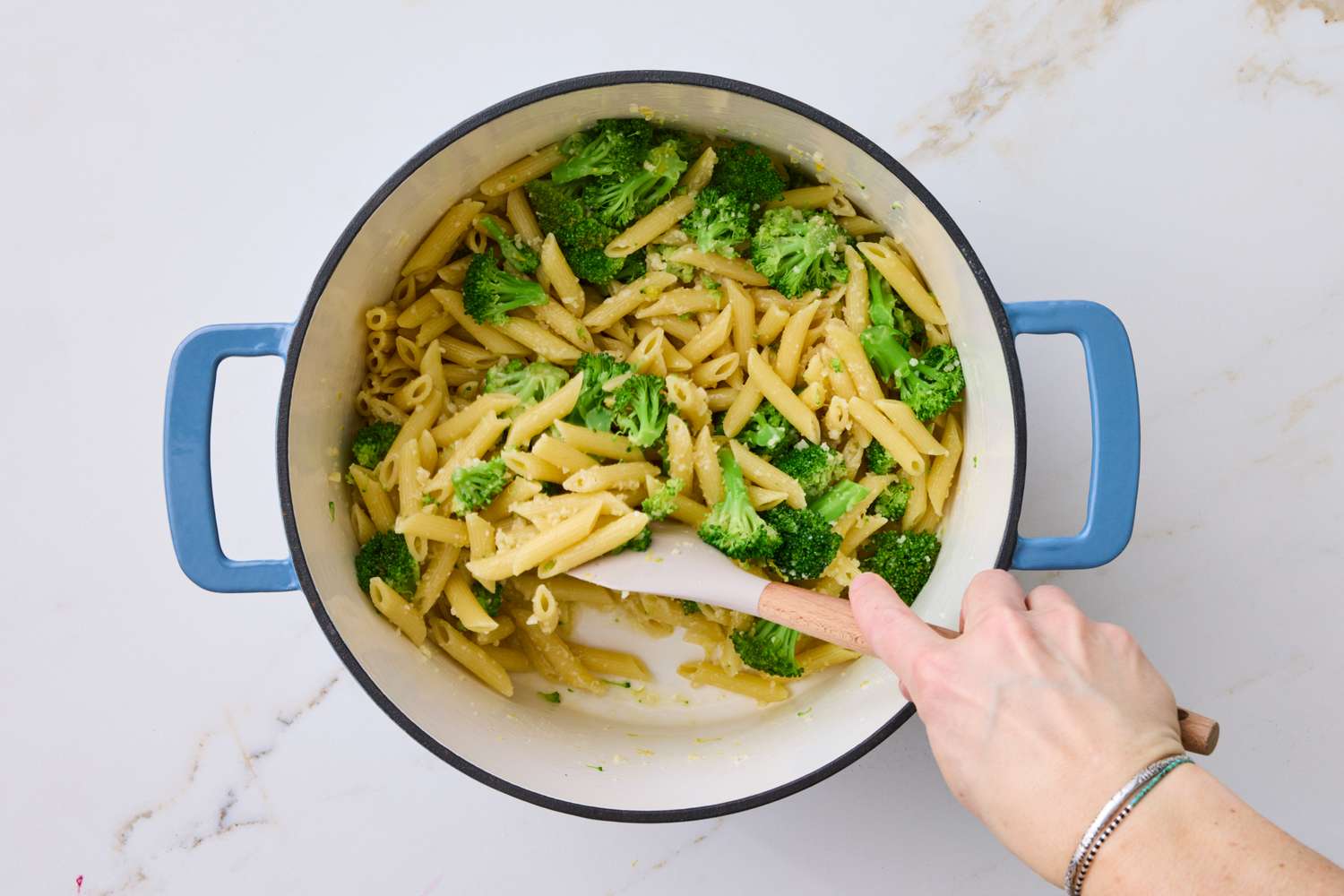 A pot of penne pasta mixed with broccoli with a hand stirring it using a spatula