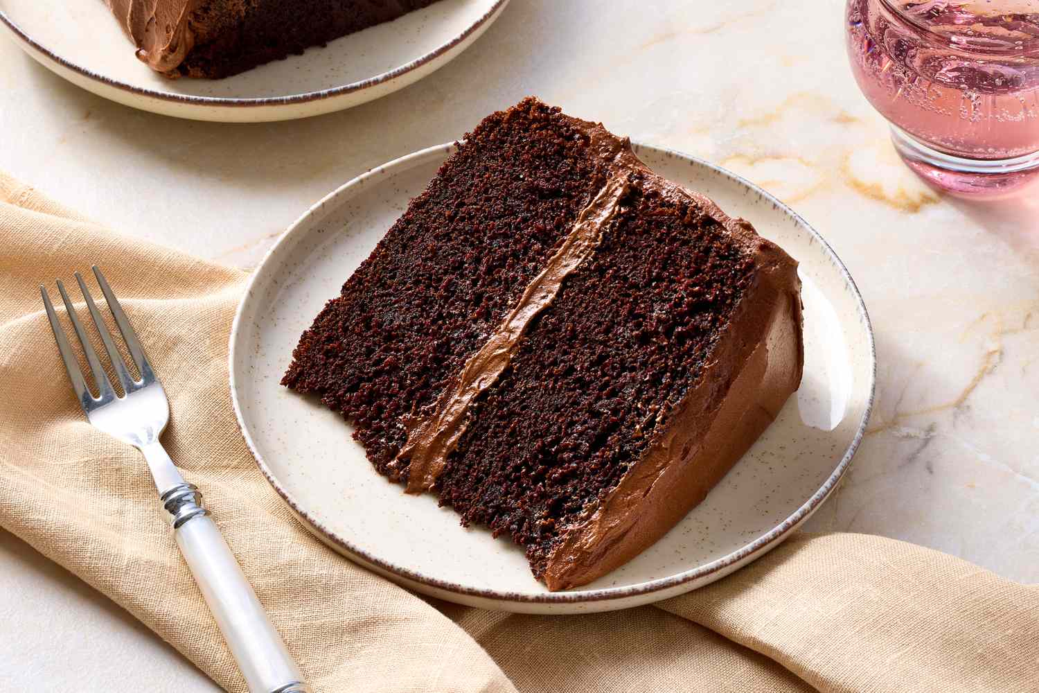 A slice of chocolate layer cake with icing on a plate accompanied by a fork and beige napkin