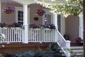 A front porch with hanging and railing flower baskets white railing and a few chairs visible
