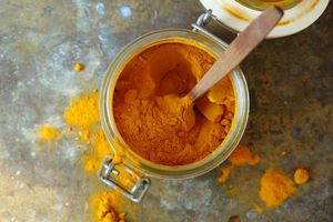 An open jar of turmeric powder with a spoon some powder spilled on a metal surface