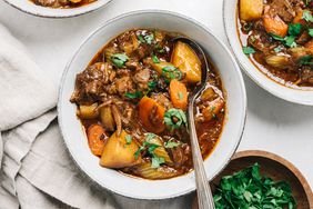 Bowls of stew with vegetables and pieces of meat, garnished with herbs, a napkin and a bowl of chopped greens beside