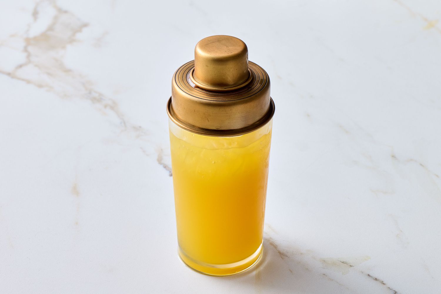 A cocktail shaker with yellow liquid on a marble surface