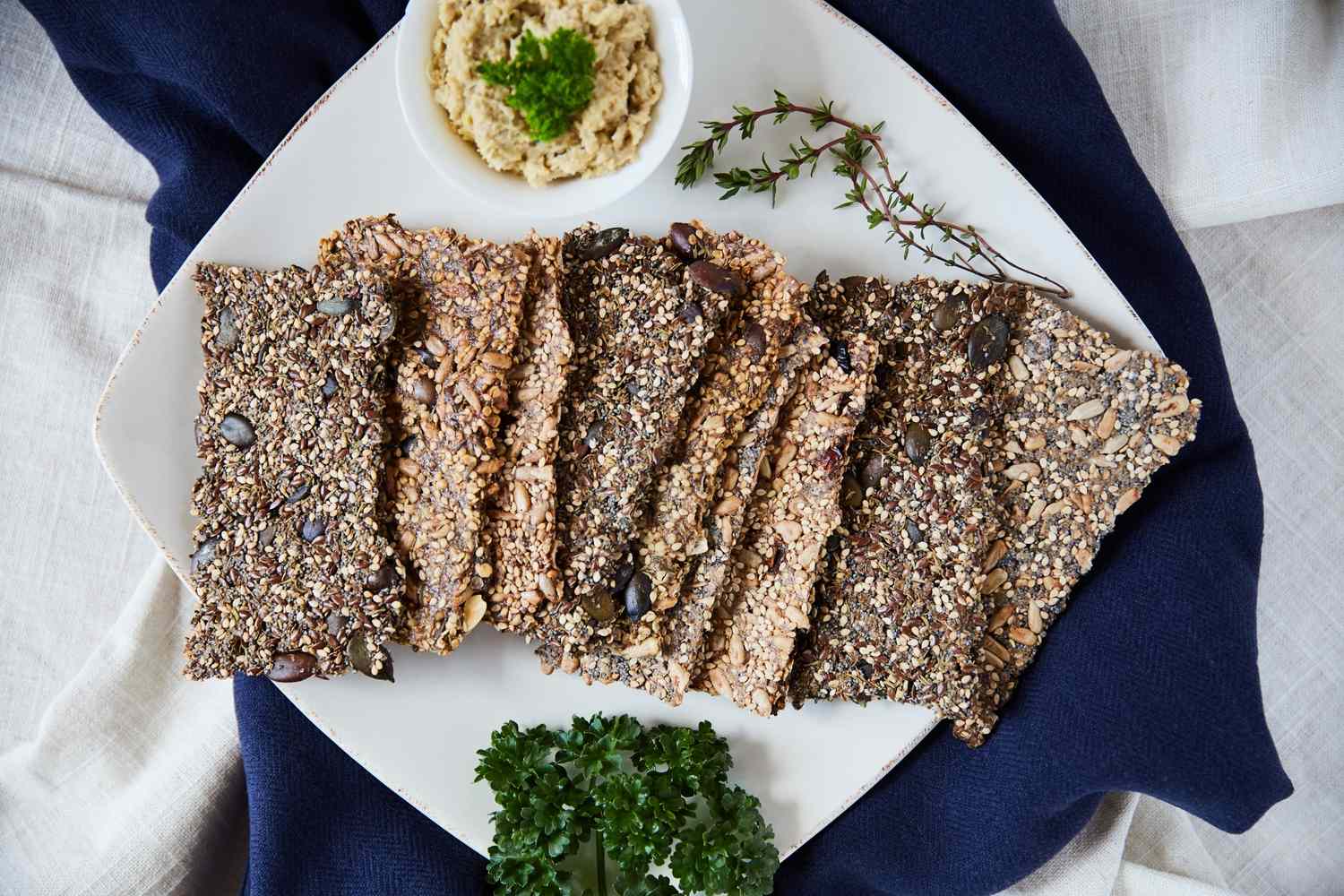 A plate with seeded crackers served next to a bowl of dip and garnished with fresh herbs