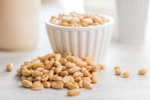 A white bowl filled with peanuts with some peanuts scattered on a surface in front of the bowl