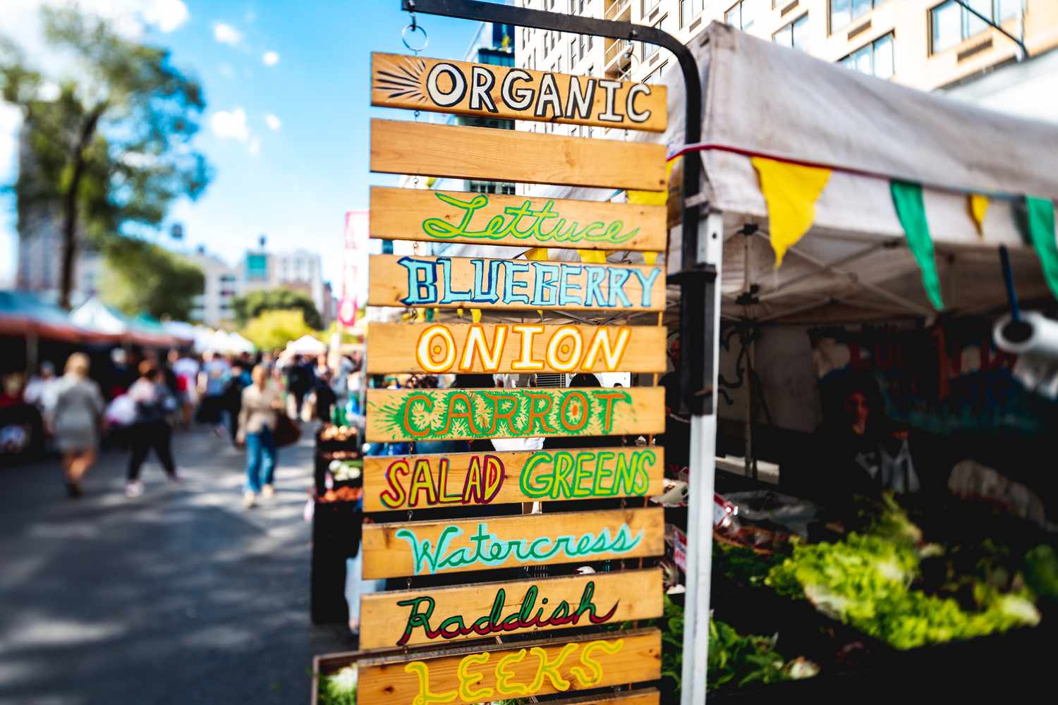 A wooden sign listing organic produce varieties at an outdoor farmers market, including lettuce, blueberry, onion, carrot, and more