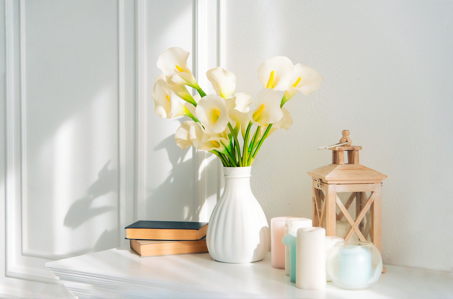 Calla lilies in a white vase on a table accompanied by books candles and a decorative lantern
