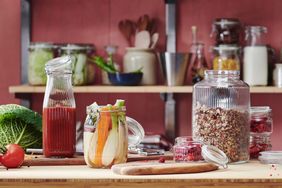 A countertop with jars of food including grains vegetables and juice with a blurred kitchen background featuring shelves and utensils