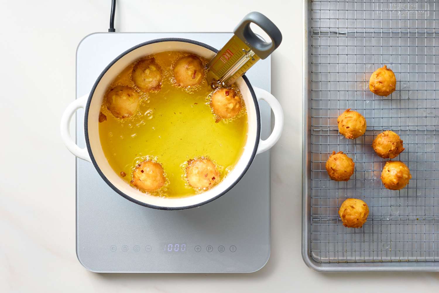 Fritters frying in oil in a pot with a thermometer next to a cooling rack with more fritters
