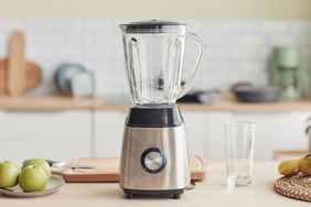A kitchen blender placed on a counter in a home kitchen setting with various items like green apples and a drinking glass nearby