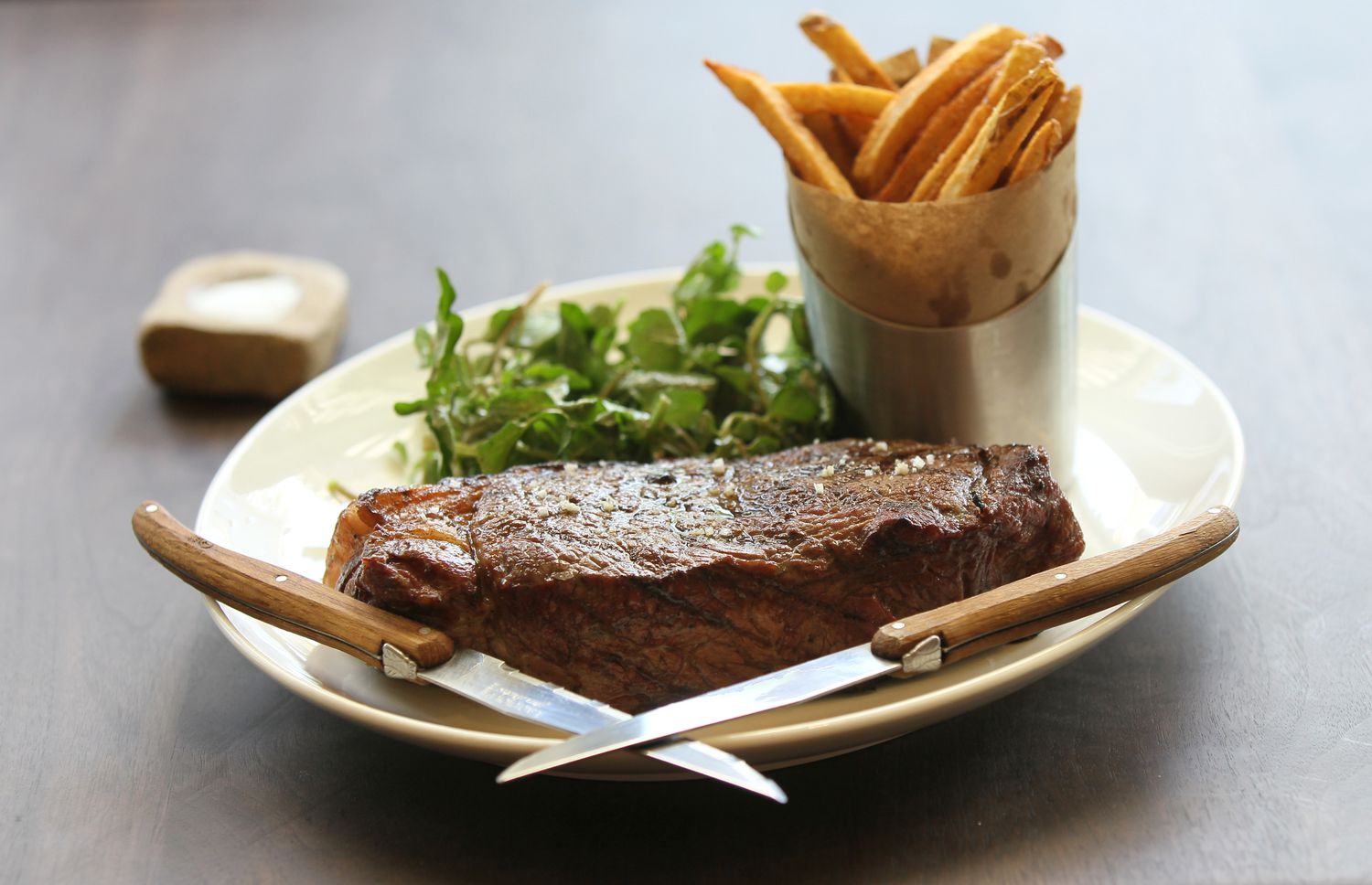 Plate containing a grilled steak, a cup of fries, and a small serving of greens