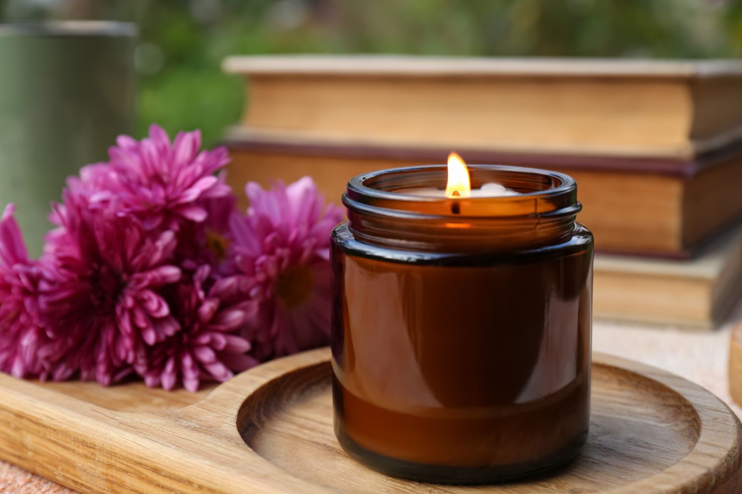 A lit candle in a glass jar placed on a wooden surface with flowers and books in the background