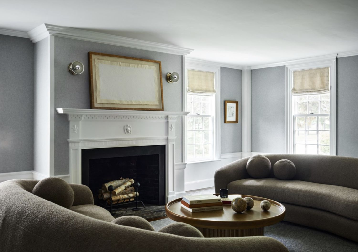 Minimalist living room by Hudson Interior Designs: taupe sculptural sofas with round pillows, white marble fireplace, wooden coffee table with books and decor, flannel wall cloth, linen curtains on windows, and natural light create a calm, stylish space.