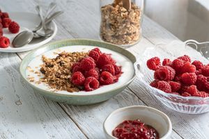 A bowl of yogurt garnished with granola and raspberries surrounded by a glass bowl of raspberries and jam on a wooden table