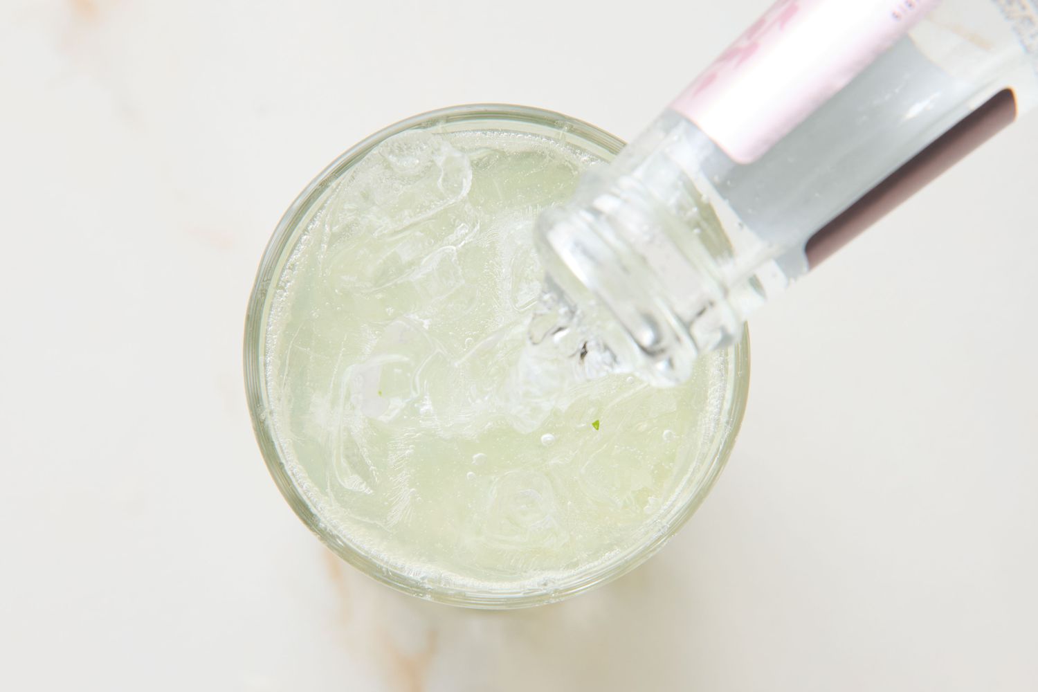 A top view of a glass filled with crushed ice and liquid being poured from a bottle, step of preparing a mojito cocktail