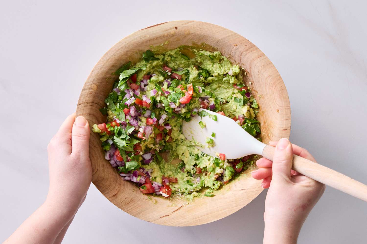 A person mixing guacamole with diced vegetables in a wooden bowl using a spatula