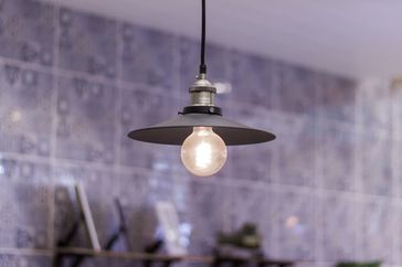 Hanging light fixture with metallic shade and exposed bulb in front of a tiled background