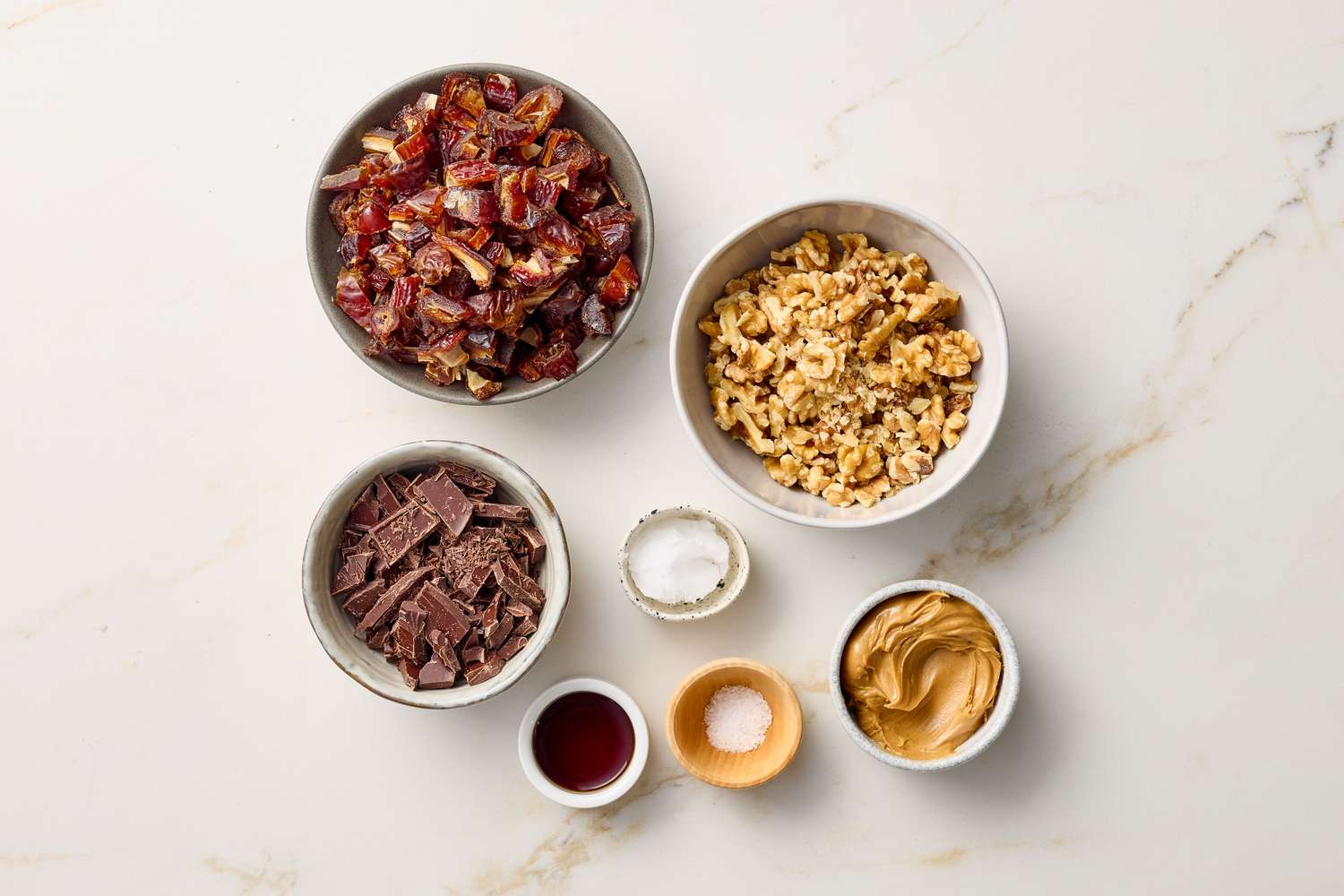Ingredients for a dessert recipe including dates walnuts chocolate vanilla extract peanut butter and salt arranged in bowls and cups