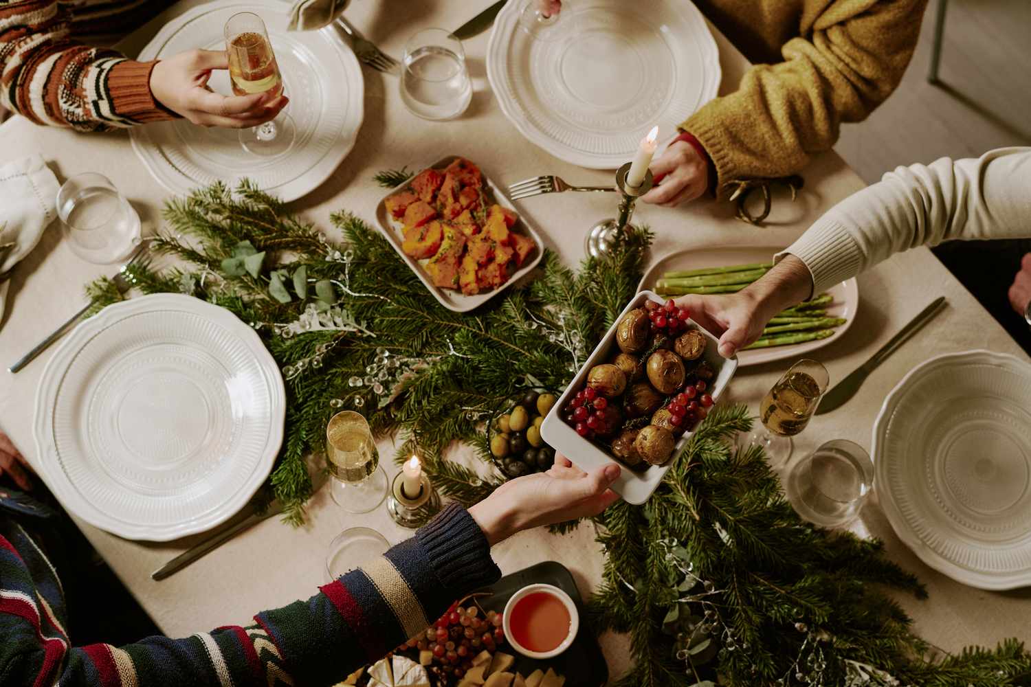 Dinner table with plates utensils and festive dishes decorated with greenery and shared by hands reaching to serve food