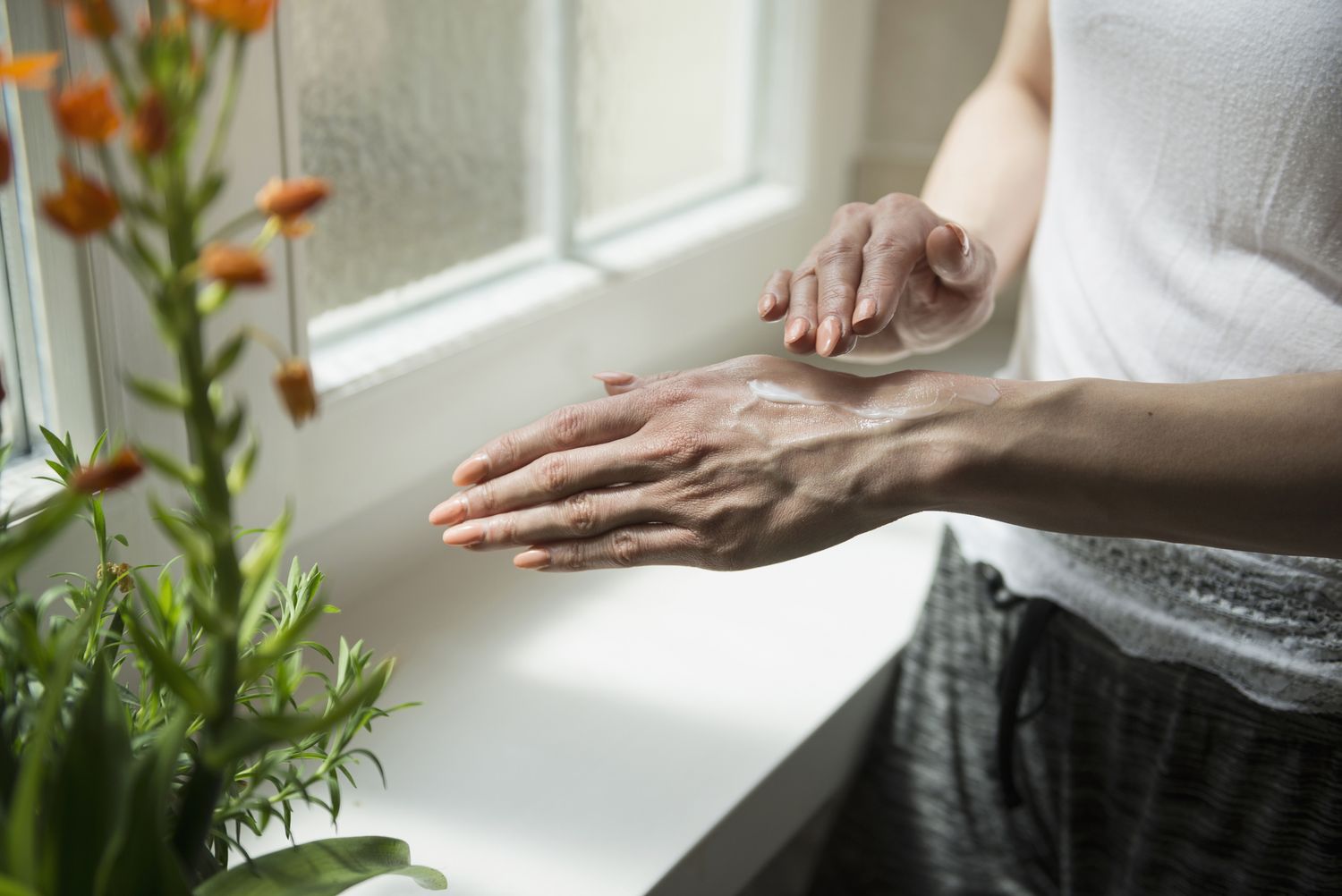 Person applying cream to their hand near a window with plants on the windowsill
