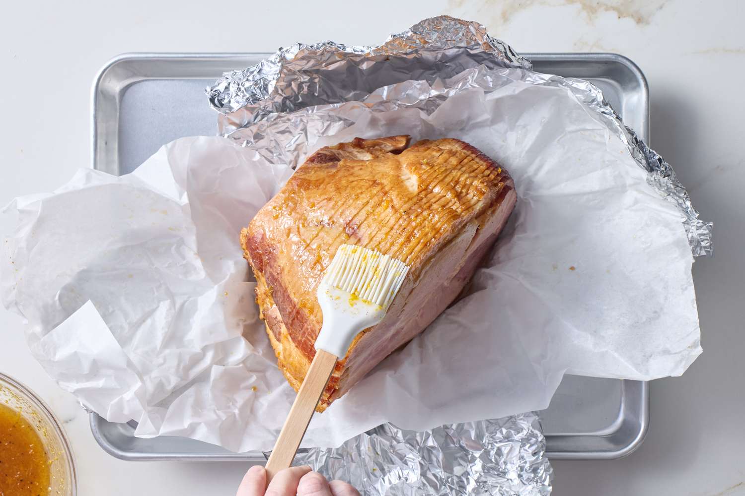 Brushing glaze onto a spiralcut ham placed on a baking sheet lined with foil and parchment paper