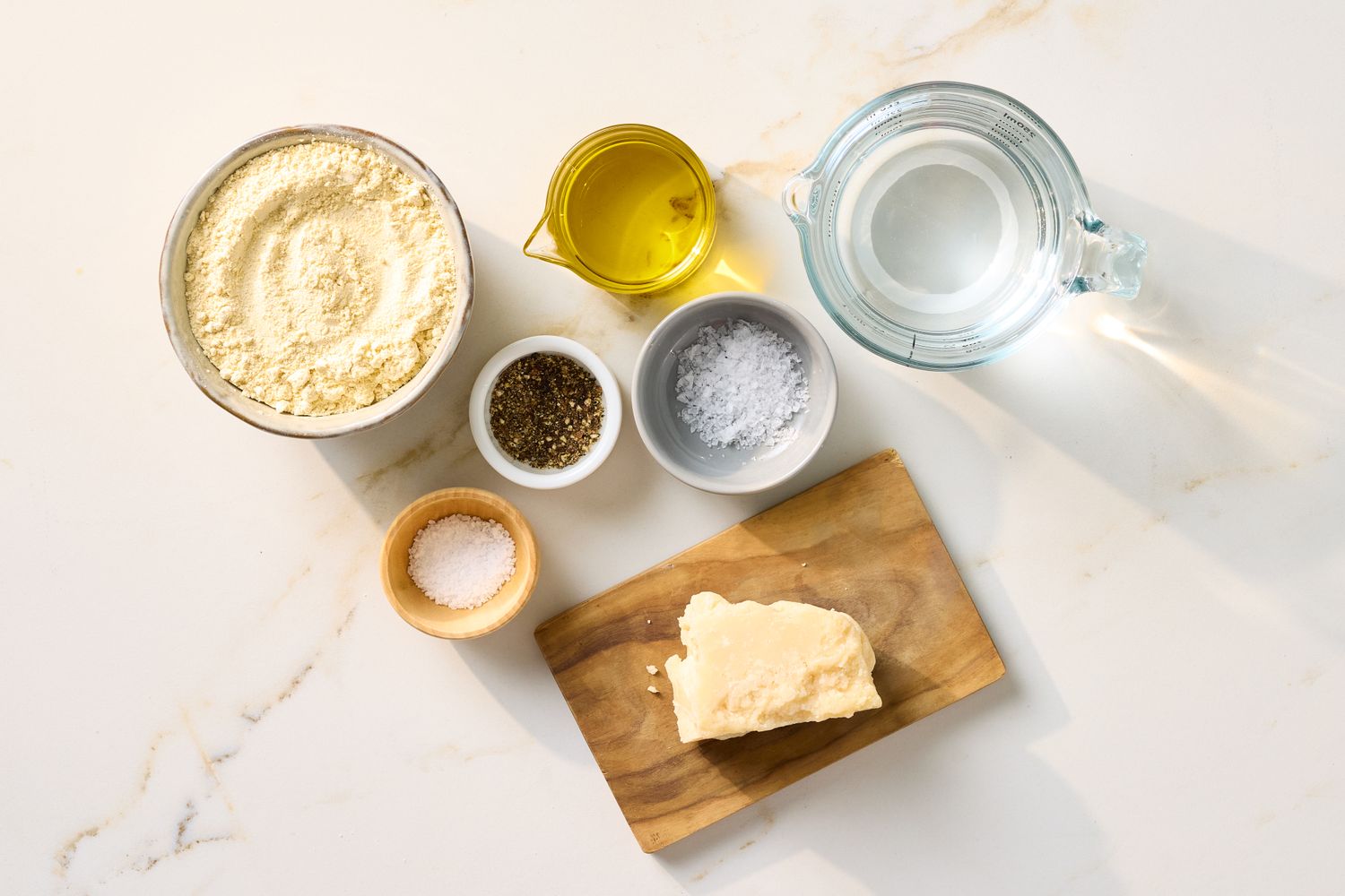 Ingredients for farinata arranged on a surface including chickpea flour olive oil water grated cheese salt and pepper