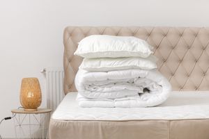 A neatly made bed with stacked bedding items including folded sheets and pillows on top a lamp on a small table nearby