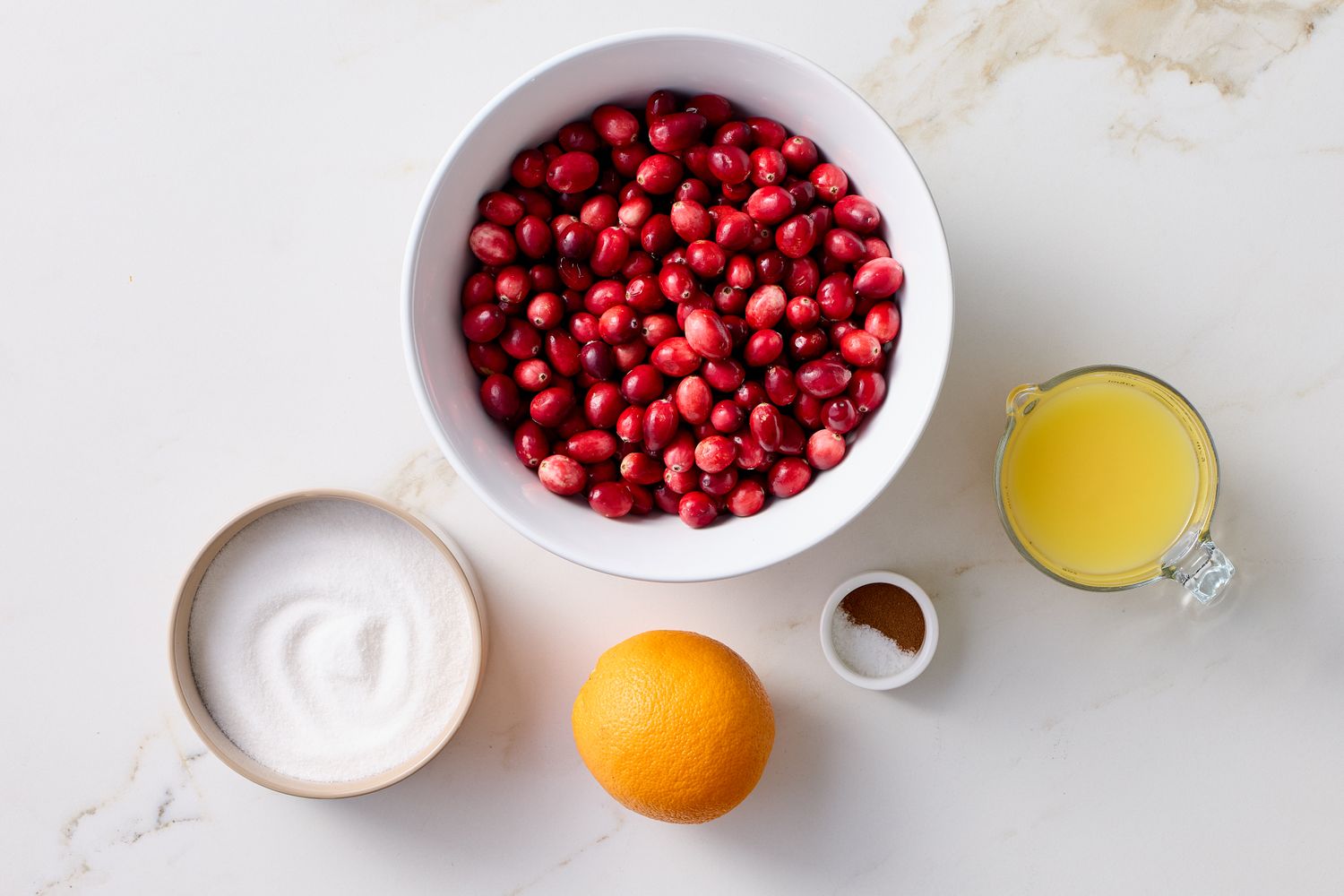 Ingredients for cranberry sauce including a bowl of cranberries sugar an orange ground cinnamon and orange juice in a cup
