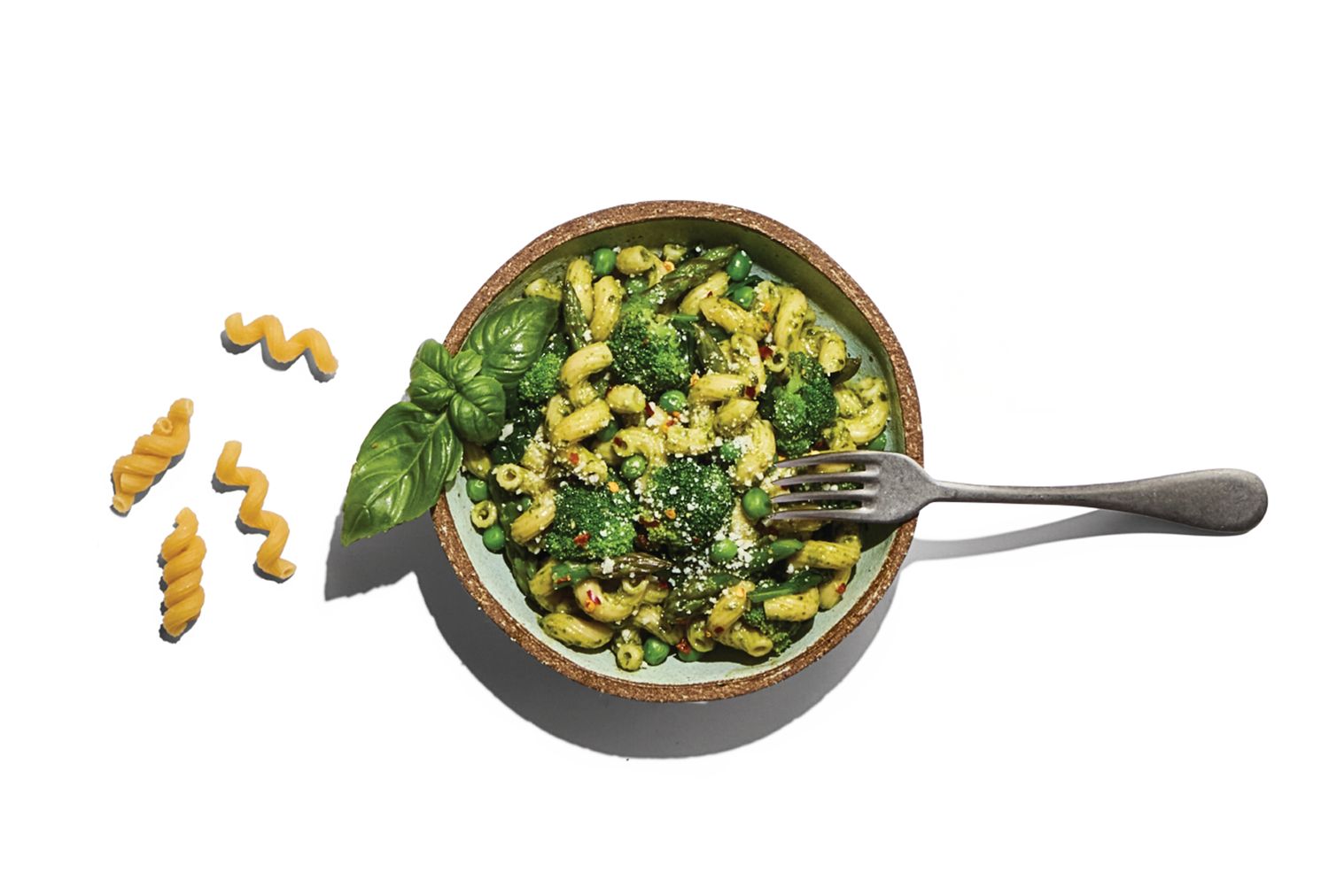 A bowl of pasta with pesto and greens garnished with basil and a fork beside it