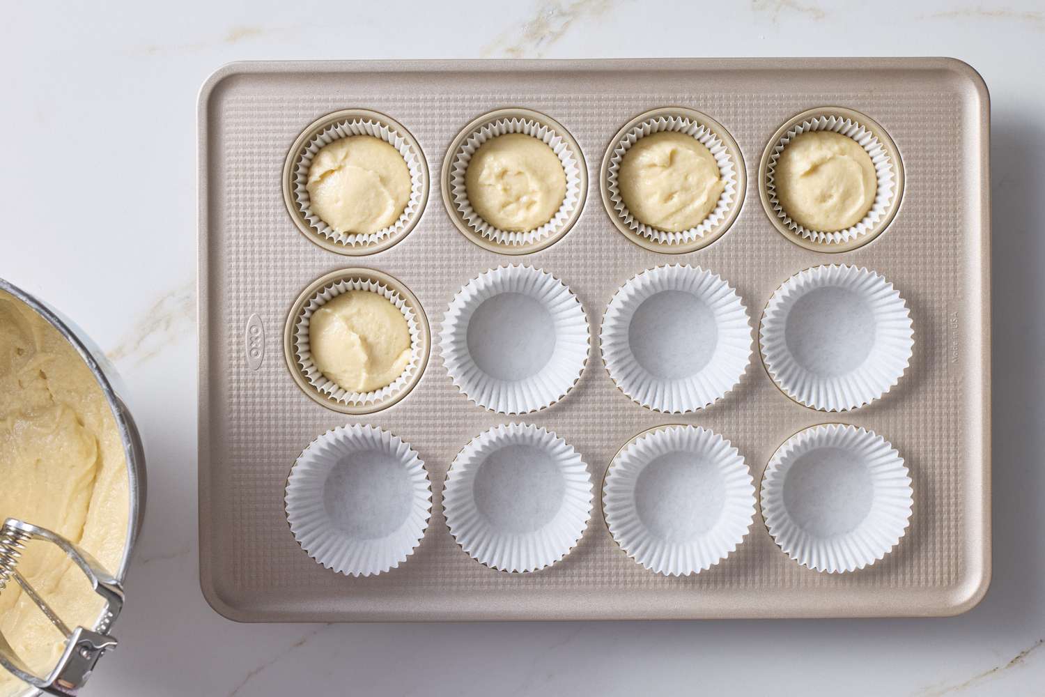 A baking tray with cupcake liners some filled with batter beside a bowl of batter and a whisk